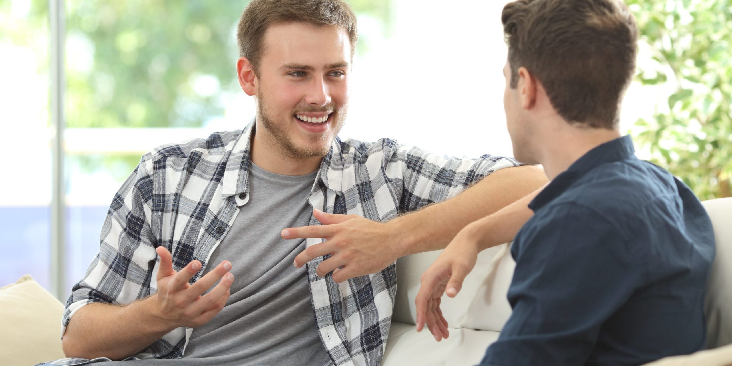 Two friends talking sitting in a couch in the living room with a window in the background at home