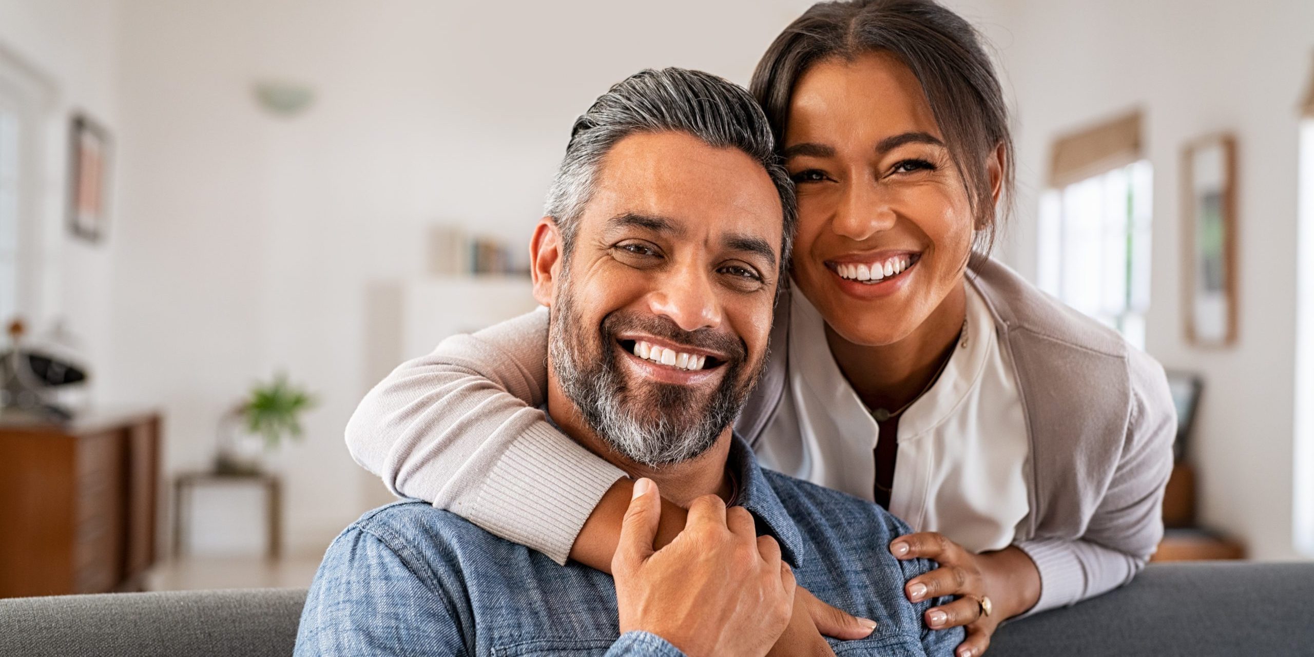 Portrait of multiethnic couple embracing and looking at camera sitting on sofa. Smiling african american woman hugging mid adult man sitting on couch from behind at home. Happy mixed race couple laugh